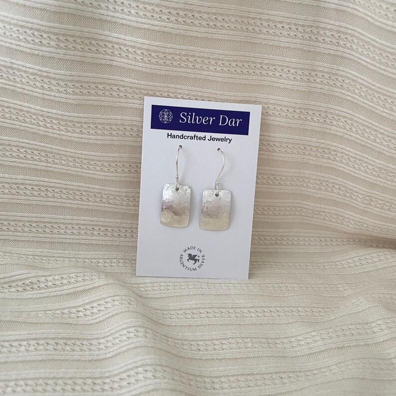 May include: A pair of silver rectangular drop earrings with a hammered texture on a white display card labeled Silver Dar Handcrafted Jewelry with a Made in Argentium Silver logo at the bottom center of the card.