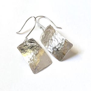 May include: A pair of silver-toned drop earrings featuring rectangular pendants with a hammered, textured surface. Each pendant is suspended from a curved silver-toned ear wire, creating a reflective and metallic appearance.