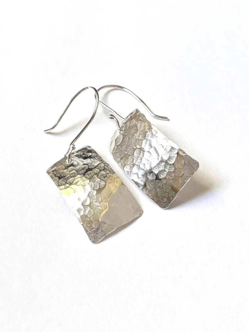 May include: A pair of silver-toned drop earrings featuring rectangular pendants with a hammered, textured surface. Each pendant is suspended from a curved silver-toned ear wire, creating a reflective and metallic appearance.