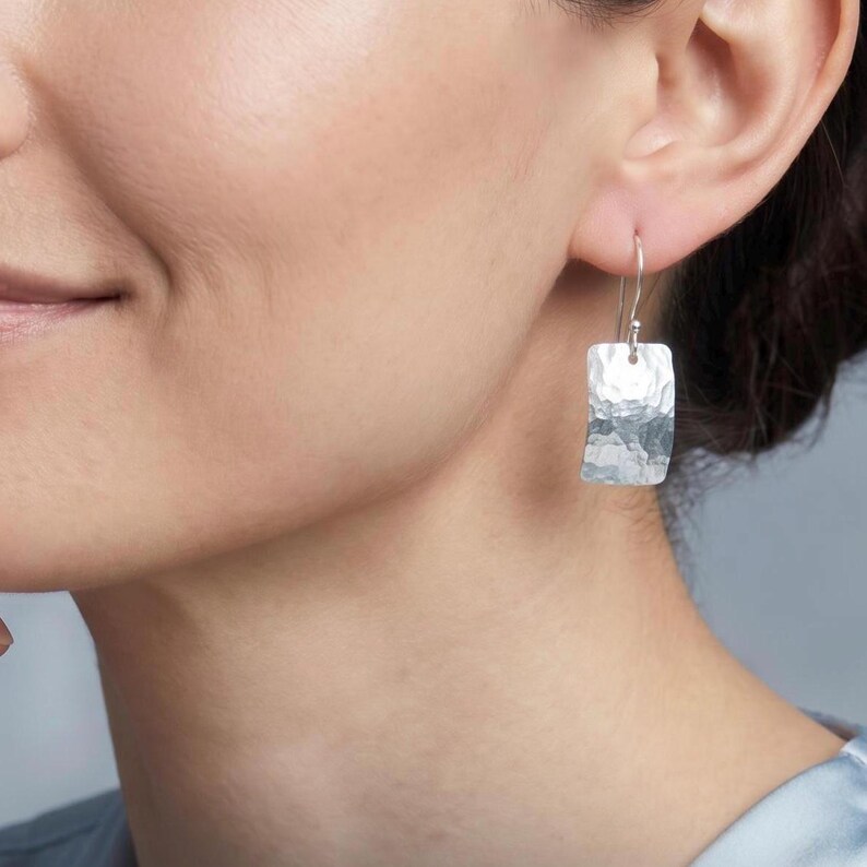 May include: A silver drop earring featuring a rectangular pendant with a textured, hammered finish. The pendant hangs from a thin, curved silver wire hook that passes through the earlobe, creating a simple and reflective metallic accessory.