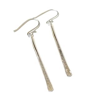 May include: A pair of long, slender silver earrings featuring a hammered, textured finish. Each earring is suspended from a curved ear wire, creating a simple, elongated drop shape with a metallic sheen that reflects light across the surface.