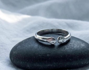 May include: A silver open-ended ring featuring a hammered texture on the band and the two rounded, slightly flattened terminals. The metal has a polished, reflective finish that catches the light, highlighting the uneven, dimpled surface.