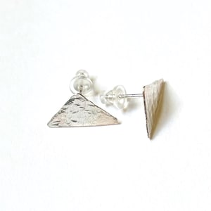 May include: A pair of silver-toned stud earrings featuring a triangular shape with a textured, hammered surface. Each earring includes a clear plastic earring back attached to a thin metal post, showcasing a metallic finish and geometric design.