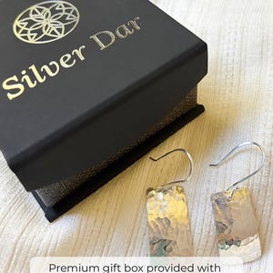 May include: A pair of silver-toned rectangular drop earrings with a hammered texture and curved ear wires, displayed next to a black jewelry box featuring the gold-colored logo and text Silver Dar on the lid.