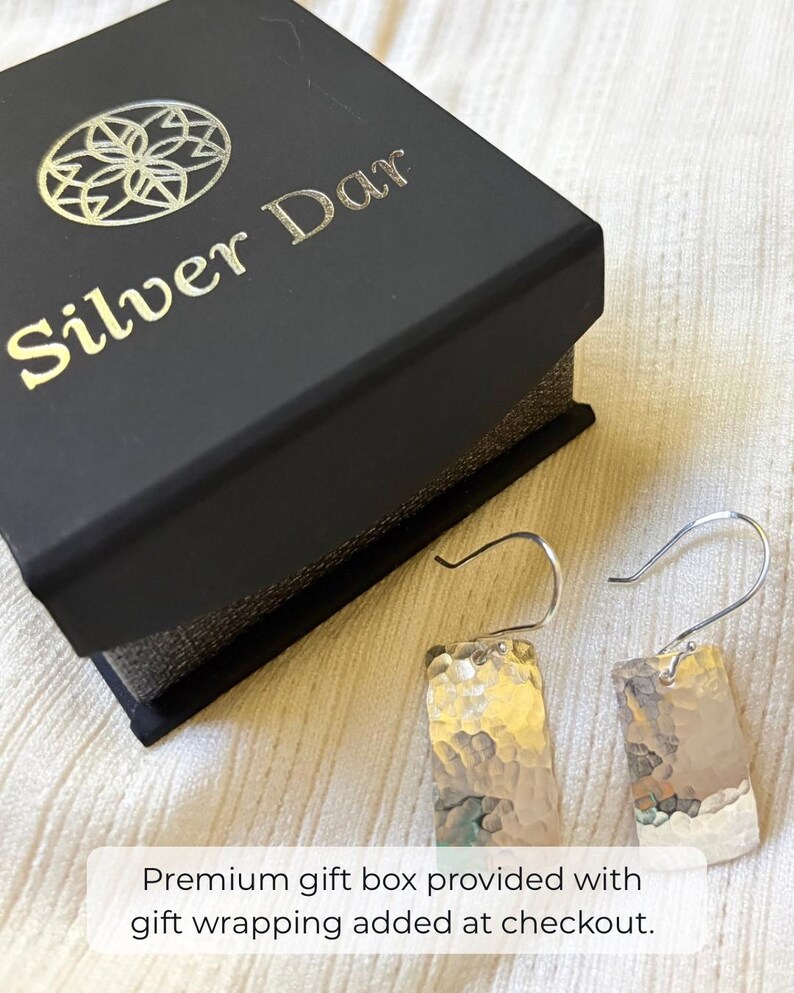 May include: A pair of silver-toned rectangular drop earrings with a hammered texture and curved ear wires, displayed next to a black jewelry box featuring the gold-colored logo and text Silver Dar on the lid.