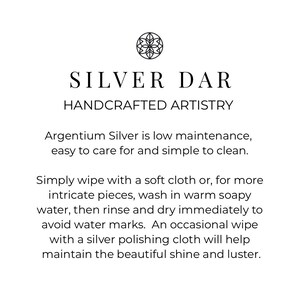 May include: A white informational card featuring a black circular logo with a floral design at the top. The text reads: SILVER DAR HANDCRAFTED ARTISTRY. Below, instructions explain that Argentium Silver is low maintenance and easy to clean by wiping with a soft cloth or washing in warm soapy water, rinsing, and drying immediately to avoid water marks.