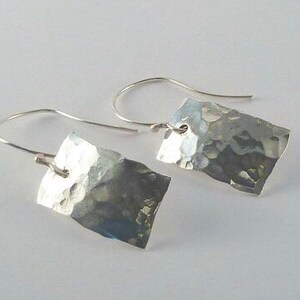 May include: A pair of silver-toned earrings featuring rectangular metal plates with a hammered, textured surface. Each plate is suspended from a curved silver-toned ear wire, creating a dangling design with reflective, uneven facets.