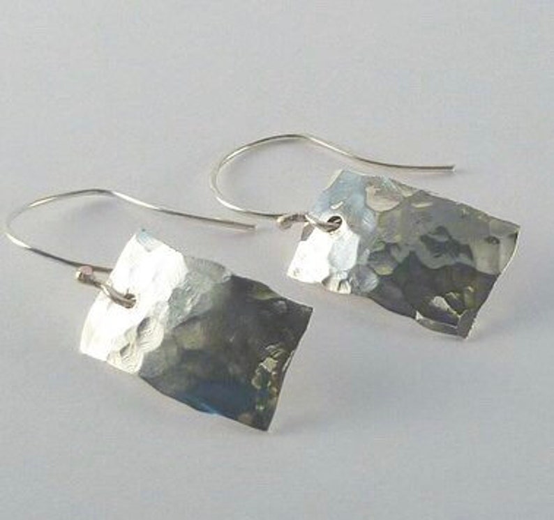 May include: A pair of silver-toned earrings featuring rectangular metal plates with a hammered, textured surface. Each plate is suspended from a curved silver-toned ear wire, creating a dangling design with reflective, uneven facets.