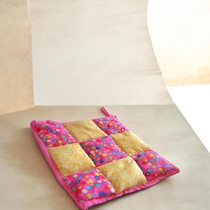 May include: A quilted hot pad with a pink border and a patchwork design. The squares alternate between a pink fabric with colorful polka dots and a yellow fabric with a floral pattern. A pink loop is attached for hanging.