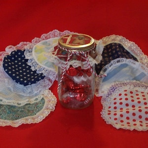 May include: A glass jar filled with red candy is surrounded by white lace and fabric circles with various patterns, including polka dots, stripes, and floral designs.