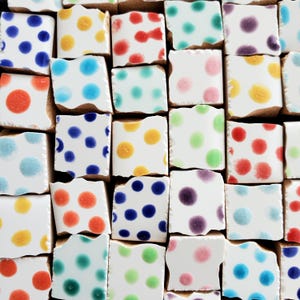May include: A collection of square ceramic tiles arranged in a grid, each featuring a white base with a single or multiple colored polka dots. The dots appear in various shades including blue, yellow, red, teal, purple, pink, and light green.