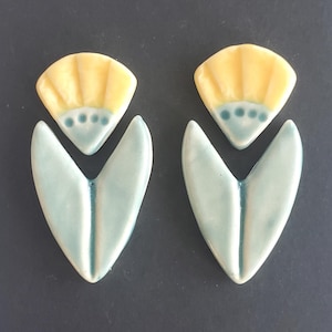 May include: Two ceramic earrings with a floral design. Each earring features a yellow, fan-shaped top with blue dots and a light blue, heart-shaped bottom. The earrings are set against a dark gray background.