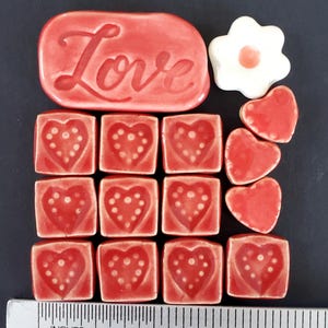 May include: A collection of red ceramic tiles and shapes. Includes a rectangular tile with the word "Love", square tiles with heart designs, a flower shape, and heart shapes. A ruler shows the objects are approximately 2 inches in length.