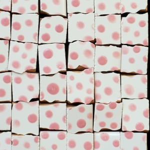 May include: A collection of small, square, white tiles with a pink polka dot pattern. The tiles are arranged in a grid, with each tile slightly irregular in shape, creating a textured surface. The pink dots vary in size.