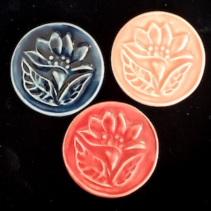 May include: Three ceramic coasters with a floral design. The coasters are round and have a raised design of a flower with leaves. The coasters are in three colors: blue, pink, and red.