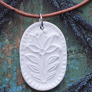May include: White ceramic pendant with a floral design, hanging on a brown leather cord. The pendant is oval shaped and has a raised floral pattern.