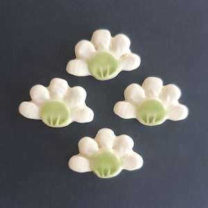 May include: Four ceramic flower-shaped embellishments. Each flower has white petals and a light green center. The embellishments are arranged on a dark blue surface. These decorative pieces could be used for crafts or jewelry making.