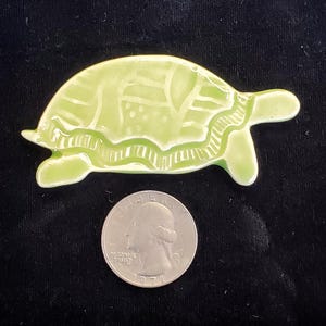 May include: A green ceramic turtle figurine with a detailed shell design. The turtle is about the size of a quarter.