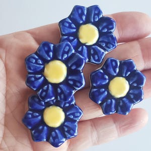 May include: A collection of four ceramic flower-shaped tiles. Each tile features a deep blue petal design surrounding a yellow center. The tiles are small, with a diameter of approximately 1 inch, and have a glossy finish.