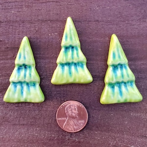 May include: Three ceramic tree-shaped ornaments with a glossy lime green and dark blue glaze finish. A copper-colored United States one-cent coin is positioned below the center tree to provide a clear sense of scale for the objects.