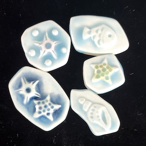 May include: A collection of six ceramic tiles with a light blue glaze. Each tile features a different embossed sea-themed design, including a fish, starfish, and seashell. The tiles are arranged on a dark surface.