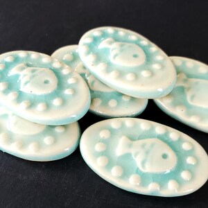 May include: A collection of oval-shaped ceramic tiles with a light blue glaze. Each tile features a white fish design in the center, surrounded by white dots. The tiles are stacked on a dark surface.
