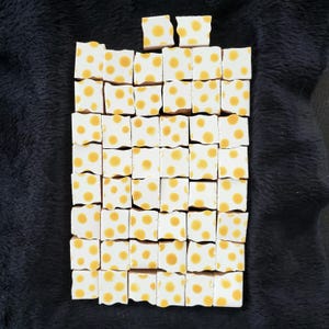 May include: A collection of small, square tiles arranged in a rectangular shape. Each tile is white with a yellow polka dot pattern. The tiles are arranged on a dark, textured surface.