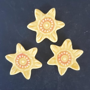 May include: Three yellow ceramic flower-shaped decorative pieces with a light orange center and a dark background. The flower petals are detailed with subtle lines. The center has a dotted pattern. These pieces could be used as coasters or decorative accents.