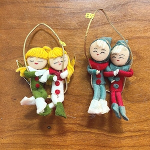 May include: Two vintage Christmas ornaments. One pair features figures in green and white outfits with yellow hair, the other pair in red and blue outfits. Each pair is suspended by a gold-colored loop.