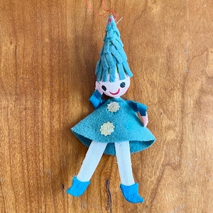 May include: A felt doll ornament featuring a teal pointed hat with fringe, a teal dress adorned with two silver star-shaped sequins, white legs, and teal boots. The doll has a round white face with black eyes and a red smile, suspended by a red string.