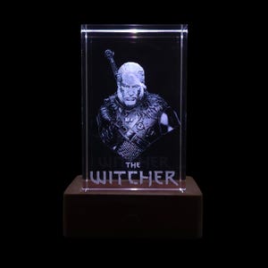 Pu&ograve; includere: Un blocco di vetro rettangolare trasparente con un ritratto inciso al laser di un personaggio dai capelli lunghi e l'elsa di una spada visibile dietro la spalla. Il testo THE WITCHER &egrave; inciso sotto la figura, su una base rettangolare.