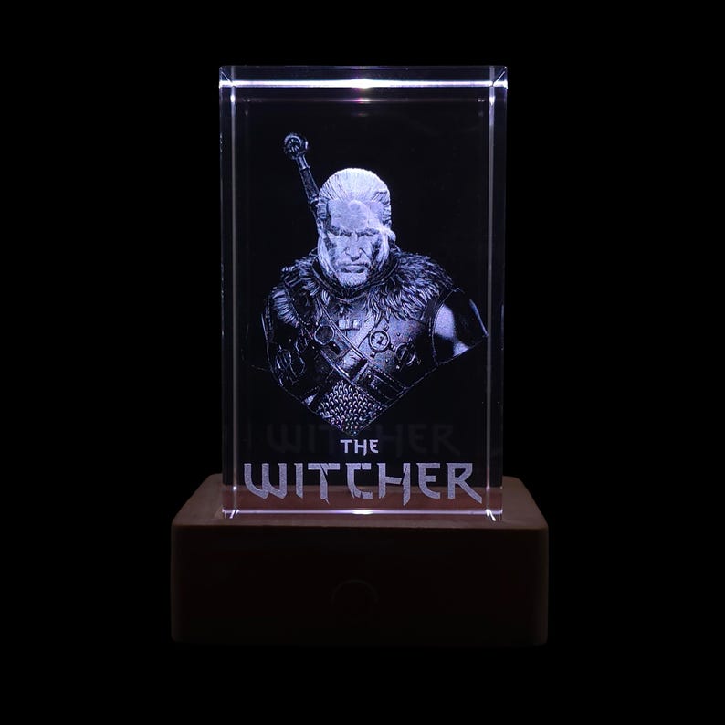 Pu&ograve; includere: Un blocco di vetro rettangolare trasparente con un ritratto inciso al laser di un personaggio dai capelli lunghi e l'elsa di una spada visibile dietro la spalla. Il testo THE WITCHER &egrave; inciso sotto la figura, su una base rettangolare.