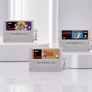 Può includere: Tre portachiavi a forma di cartuccia di gioco Super Nintendo grigio chiaro. In alto a sinistra Battletoads in Battlemaniacs, in alto a destra Super Mario World e in basso al centro The Legend of Zelda: A Link to the Past.