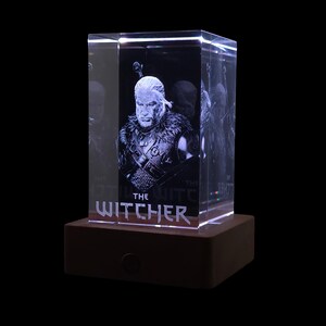 Pu&ograve; includere: Un blocco di vetro rettangolare trasparente con un ritratto 3D inciso al laser di un personaggio con capelli lunghi e una tunica con colletto di pelliccia. Sotto la figura, il testo THE WITCHER &egrave; inciso nel vetro su una base solida.