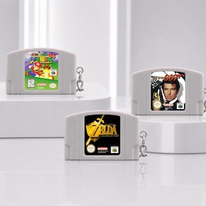 May include: Three grey plastic video game cartridge keychains. One features the Super Mario 64 label, one displays The Legend of Zelda Ocarina of Time artwork, and the third shows the 007 GoldenEye game cover with a portrait of Pierce Brosnan.