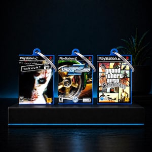 Könnte beinhalten: Drei PlayStation 2-Spiel-Schlüsselanhänger. Die Schlüsselanhänger zeigen die Spielcover von "Manhunt", "Need for Speed Underground 2" und "Grand Theft Auto: San Andreas". Jeder Schlüsselanhänger hat einen blauen Rand und einen silbernen Ring.