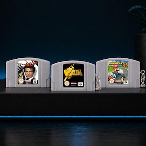 May include: Three grey Nintendo 64 game cartridge keyrings. The keyrings feature the game titles "007", "Zelda", and "Mario Kart". The "007" keyring has a picture of Pierce Brosnan. The "Zelda" keyring has a gold logo. The "Mario Kart" keyring has a picture of Mario.