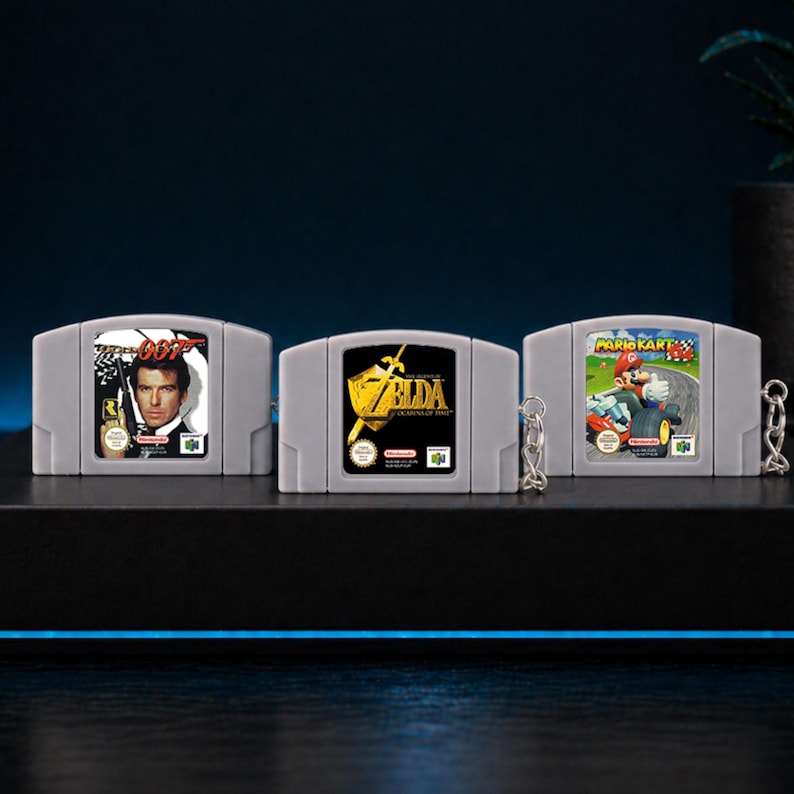 May include: Three grey Nintendo 64 game cartridge keyrings. The keyrings feature the game titles "007", "Zelda", and "Mario Kart". The "007" keyring has a picture of Pierce Brosnan. The "Zelda" keyring has a gold logo. The "Mario Kart" keyring has a picture of Mario.