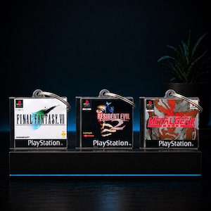 以下が含まれることがあります： 3つのPlayStationゲームケースキーホルダー。「ファイナルファンタジーVII」、「バイオハザード2」、「メタルギアソリッド」のゲームタイトルが特徴です。各キーホルダーにはシルバーのキーリングが付いており、黒いスタンドに展示されています。