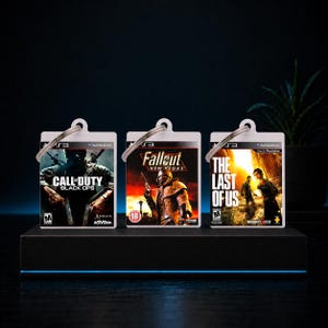 Könnte beinhalten: Drei Schlüsselanhänger mit Videospiel-Hüllen. Die Schlüsselanhänger zeigen die Spieletitel "Call of Duty: Black Ops", "Fallout: New Vegas" und "The Last of Us". Jeder Anhänger hat einen silbernen Schlüsselring.