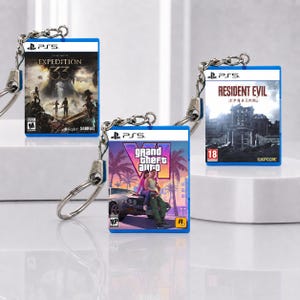 May include: Three miniature rectangular keychains featuring PlayStation 5 game case art for Expedition 33, Grand Theft Auto VI, and Resident Evil Requiem. Each keychain has a silver metal chain and ring attached to a blue plastic frame.