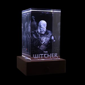 Pu&ograve; includere: Un blocco di vetro rettangolare trasparente con un ritratto inciso al laser di un personaggio dai capelli lunghi con una spada sulla schiena. Il testo THE WITCHER &egrave; inciso in basso. Il blocco poggia su una base rettangolare in legno marrone.