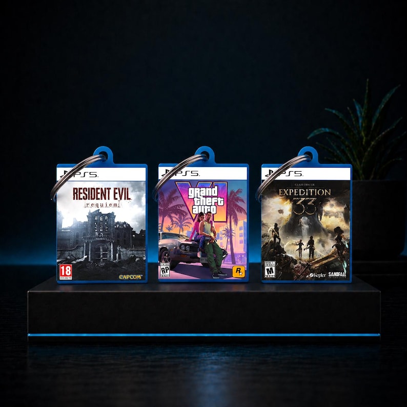 Może przedstawiać: Trzy breloczki do gier PS5. Breloczki przedstawiają tytuły gier "Resident Evil Requiem", "Grand Theft Auto V" i "Expedition 33". Każdy breloczek ma niebieską obw&oacute;dkę i srebrny pierścień.