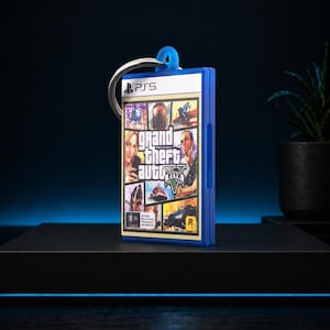 Może przedstawiać: Niebieski brelok do kluczy z grafiką z okładki gry Grand Theft Auto V. Brelok ma srebrny pierścień i niebieską pętlę. Logo gry i postacie są widoczne z przodu. Logo PS5 znajduje się na g&oacute;rze.