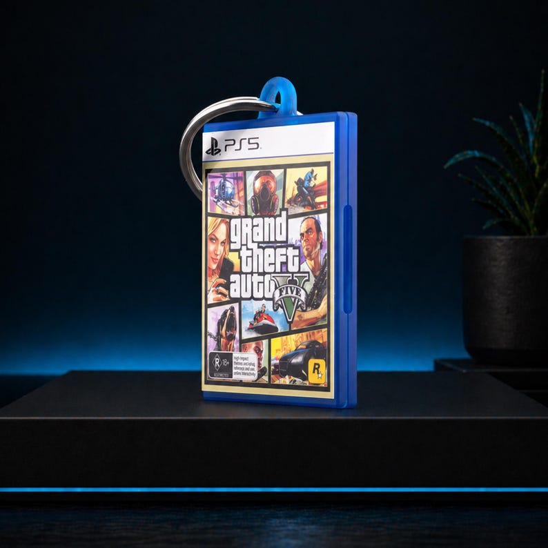 Może przedstawiać: Niebieski brelok do kluczy z grafiką z okładki gry Grand Theft Auto V. Brelok ma srebrny pierścień i niebieską pętlę. Logo gry i postacie są widoczne z przodu. Logo PS5 znajduje się na g&oacute;rze.
