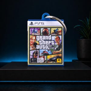 Może przedstawiać: Brelok z okładką gry Grand Theft Auto V. Brelok ma niebieską obw&oacute;dkę i srebrny k&oacute;łko na klucze. Okładka gry przedstawia r&oacute;żne postacie i sceny z gry, z logo PlayStation 5 u g&oacute;ry.