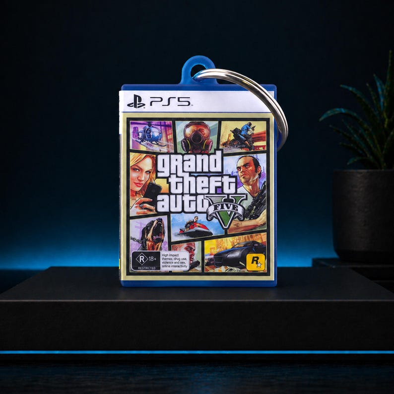 Może przedstawiać: Brelok z okładką gry Grand Theft Auto V. Brelok ma niebieską obw&oacute;dkę i srebrny k&oacute;łko na klucze. Okładka gry przedstawia r&oacute;żne postacie i sceny z gry, z logo PlayStation 5 u g&oacute;ry.