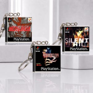 Könnte beinhalten: Drei quadratische Schlüsselanhänger mit Miniatur-PlayStation-Spielhüllen für Metal Gear Solid, Silent Hill und Resident Evil 2. Jeder Schlüsselanhänger verfügt über eine silberne Metallkette und einen Ring und zeigt das Original-Cover-Art.
