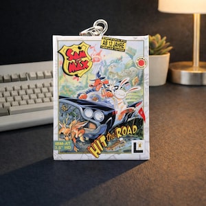 May include: A Sam & Max "Hit the Road" keychain featuring a colorful cartoon image of the game's characters in a car. The keychain has a silver-colored metal ring at the top. The game's title and logo are prominently displayed.