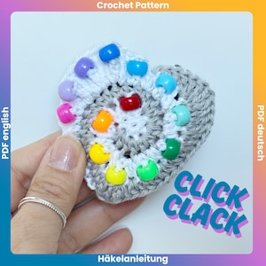 May include: A heart-shaped crochet piece featuring a gray center, a white border, and colorful plastic pony beads in red, orange, yellow, green, light blue, purple, and pink. The text CLICK CLACK is printed in blue and purple on the lower right side.